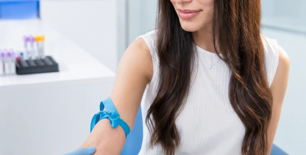 Una persona sentada con el brazo extendido y una banda elástica azul en el antebrazo, con una gradilla de tubos de muestra al fondo